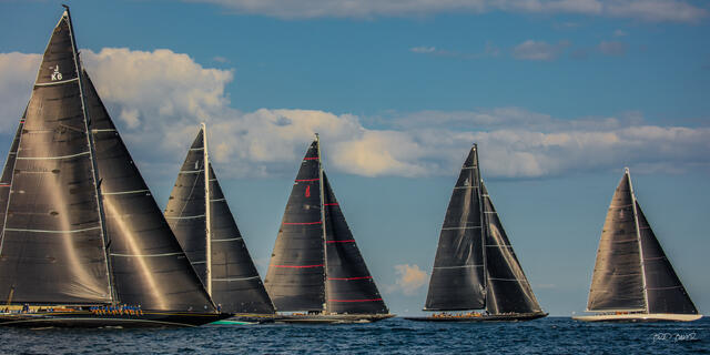 Fleet of 5 towering black-sailed J Class yachts racing tightly together under a partly cloudy sky