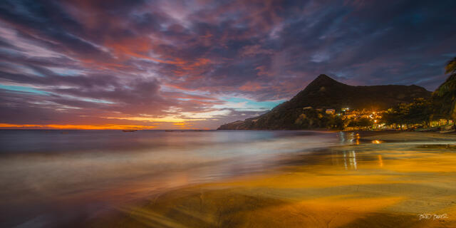 Panoramic long-exposure twilight over a calm beach with a glowing hilltop village and vivid clouds in Martinique