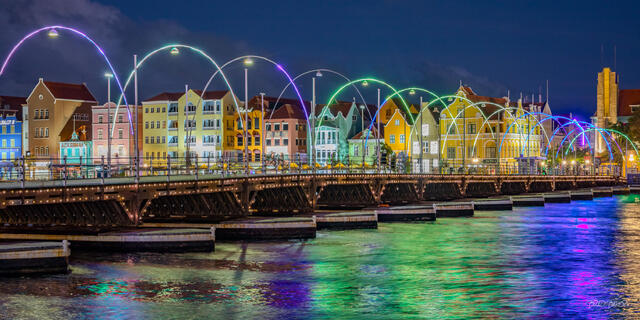 Nighttime waterfront cityscape with colorful illuminated arches, historic buildings, and vibrant reflections across the water at Willemstad Curacao