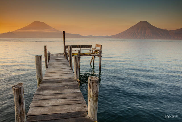 Rustic lakeside dock at dramatic sunrise, twin volcanoes silhouetted against orange sky, Lake Atitlán Guatemala