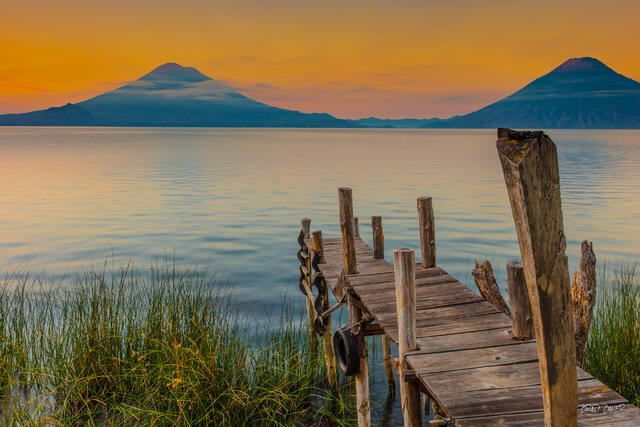 Decaying wooden dock at vivid orange sunrise with twin volcanic peaks reflected in still water, Guatemala