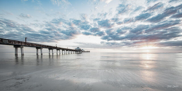 Panorama of a long pier at Fort Myers Beach extending into calm ocean under a pastel bright white sky with soft clouds and a dramatic sunburst at sunset