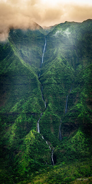 Twin waterfalls cascading down a steep green Kauai mountainside beneath golden misty clouds.