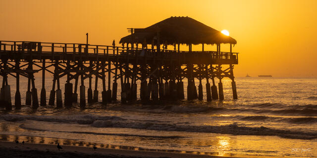 Silhouetted pier at Cocoa Beach Florida with thatched-roof tiki bar at sunrise, golden sun glowing behind the structure over the ocean
