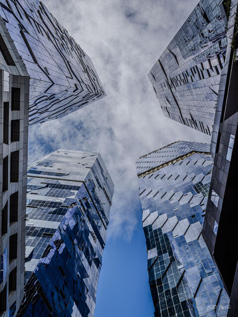Glass skyscrapers converge above against a cloudy blue sky in an upward-looking architectural photograph in Melbourne