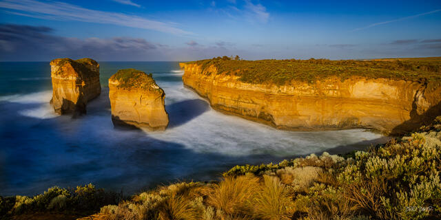 Golden sea stacks and rugged cliffs rise above smooth ocean waves at sunrise along the Australian coast
