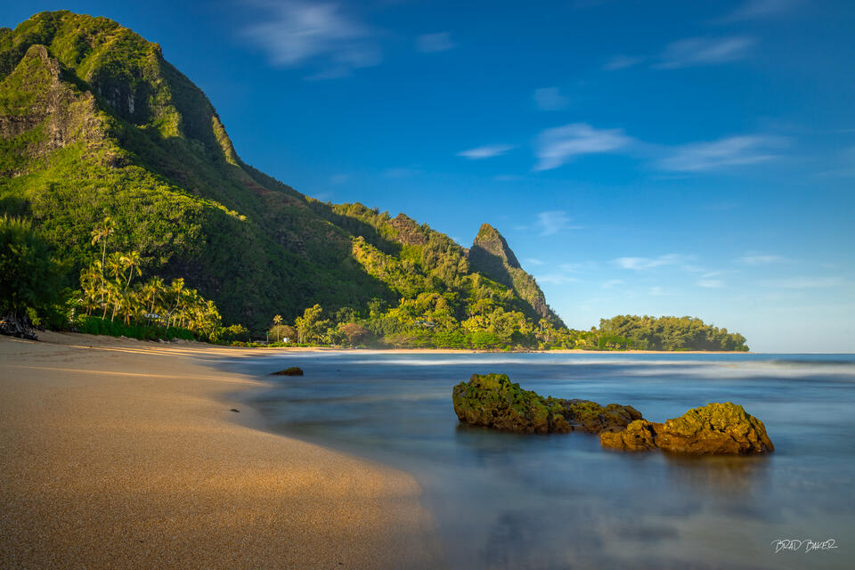 Sunlit green cliffs and Mount Makana rise above a quiet sandy beach at Tunnels, Kauai