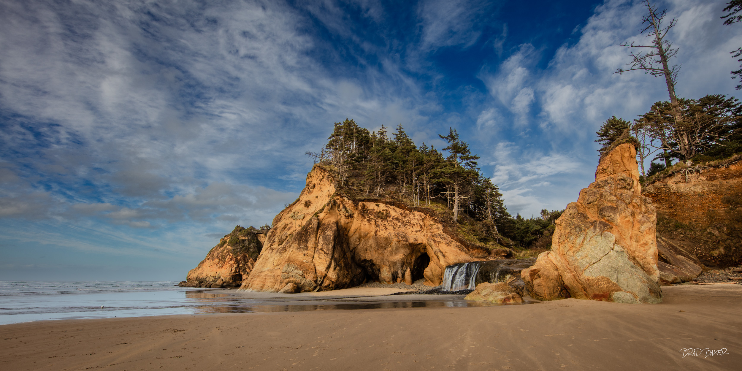 FOREST MEETS THE SEA Panorama of a distant waterfall cascading down a sandstone wall onto a sandy beach under a deep blue sky with some clouds at Hug Point Oregon