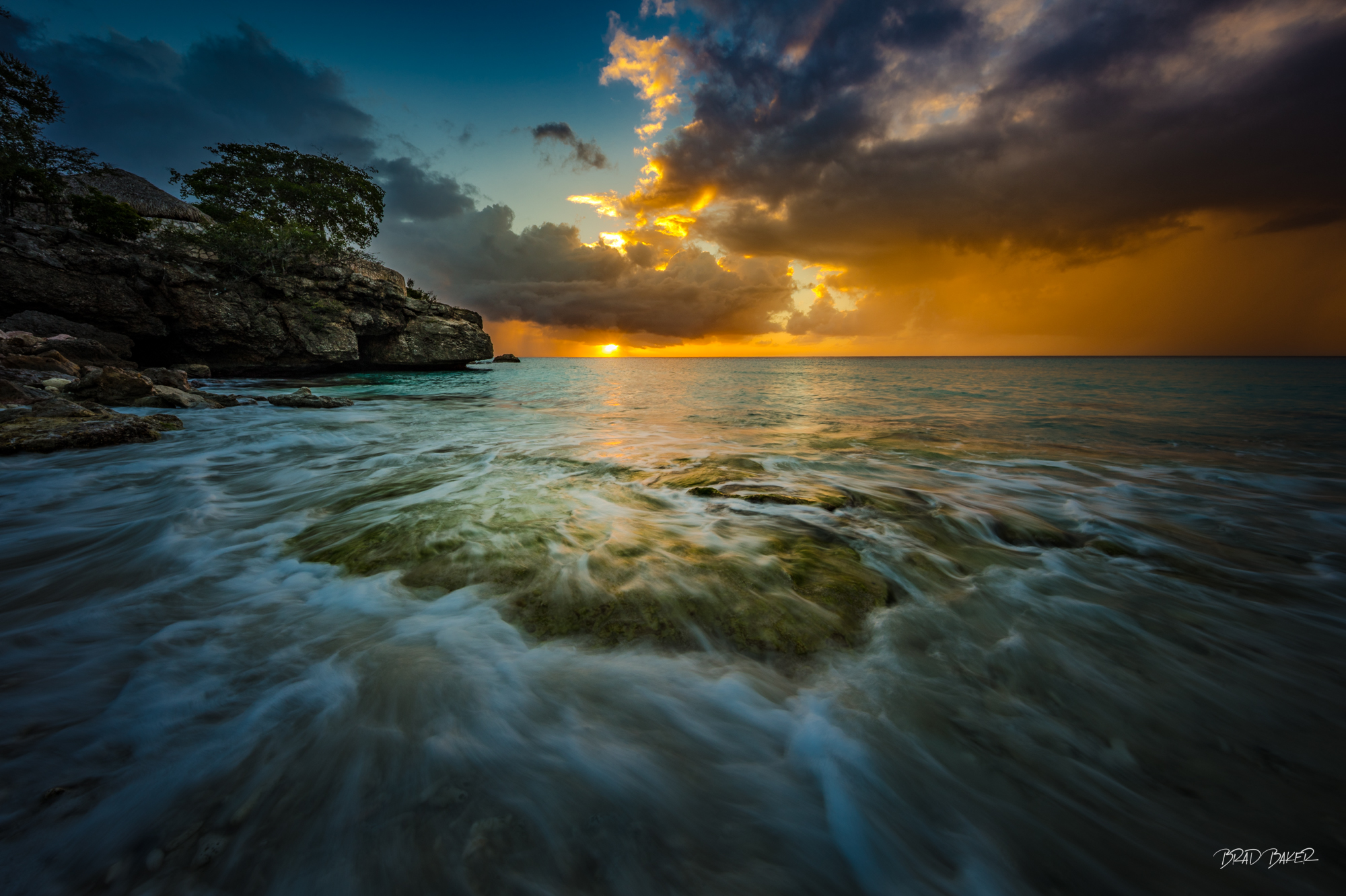 ETHERAL SHORE Dramatic tropical sunset over a rocky shoreline with flowing water and illuminated clouds in Curacao