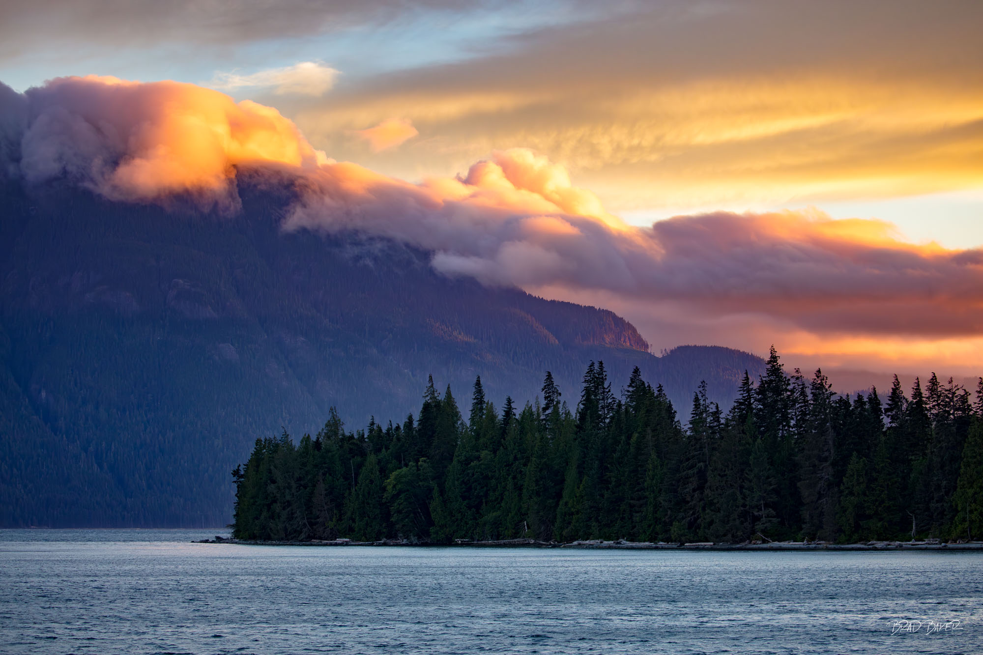 FIRE IN THE SKY Dramatic fiery sunset clouds rolling over dark coastal mountain range above forested island and calm inlet in Johnstone Strait British Columbia