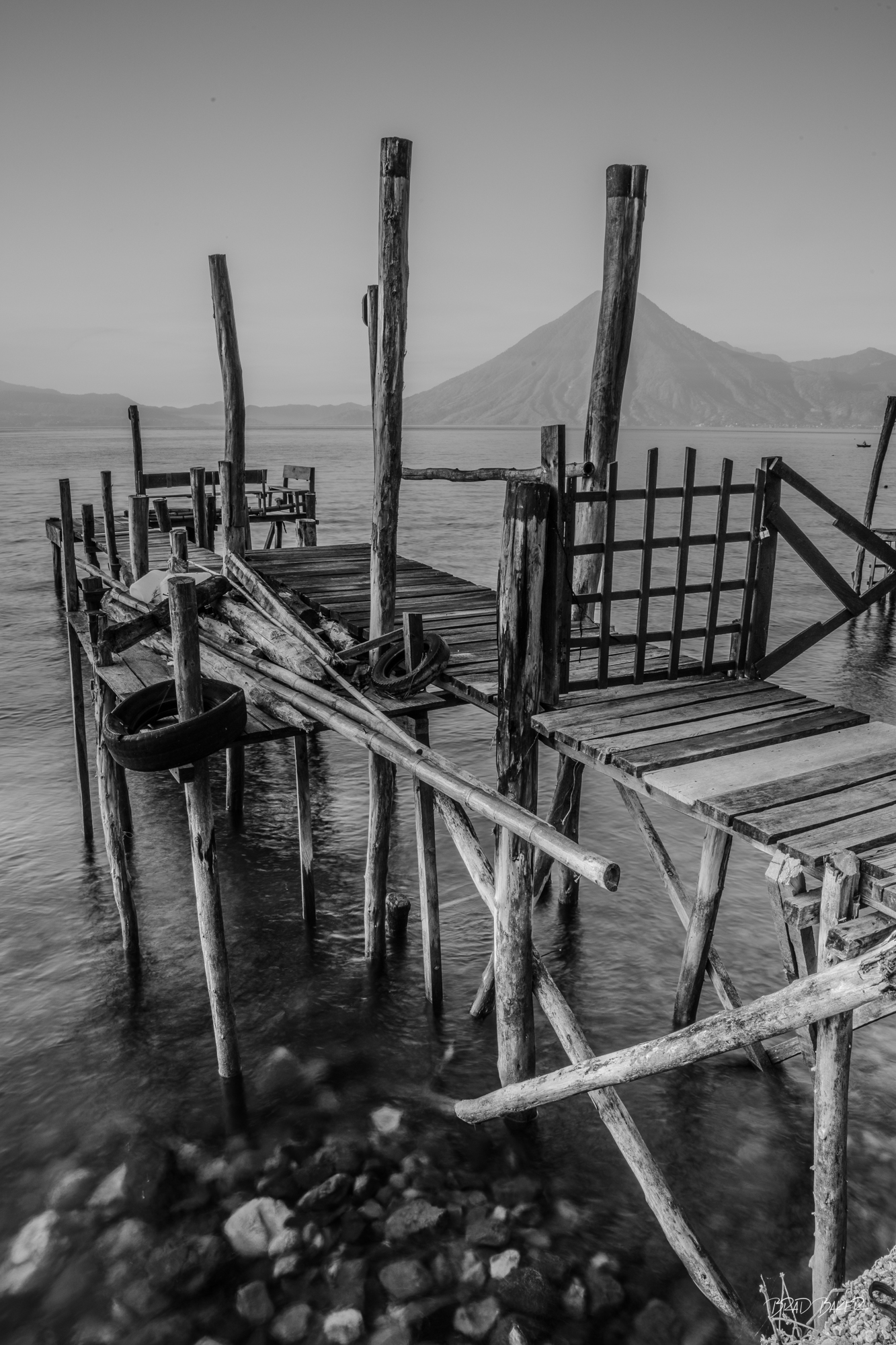 Built from the land Black and white vertical orientation of a rustic wooden dock over Lake Atitlan Guatemala with volcano in the distance