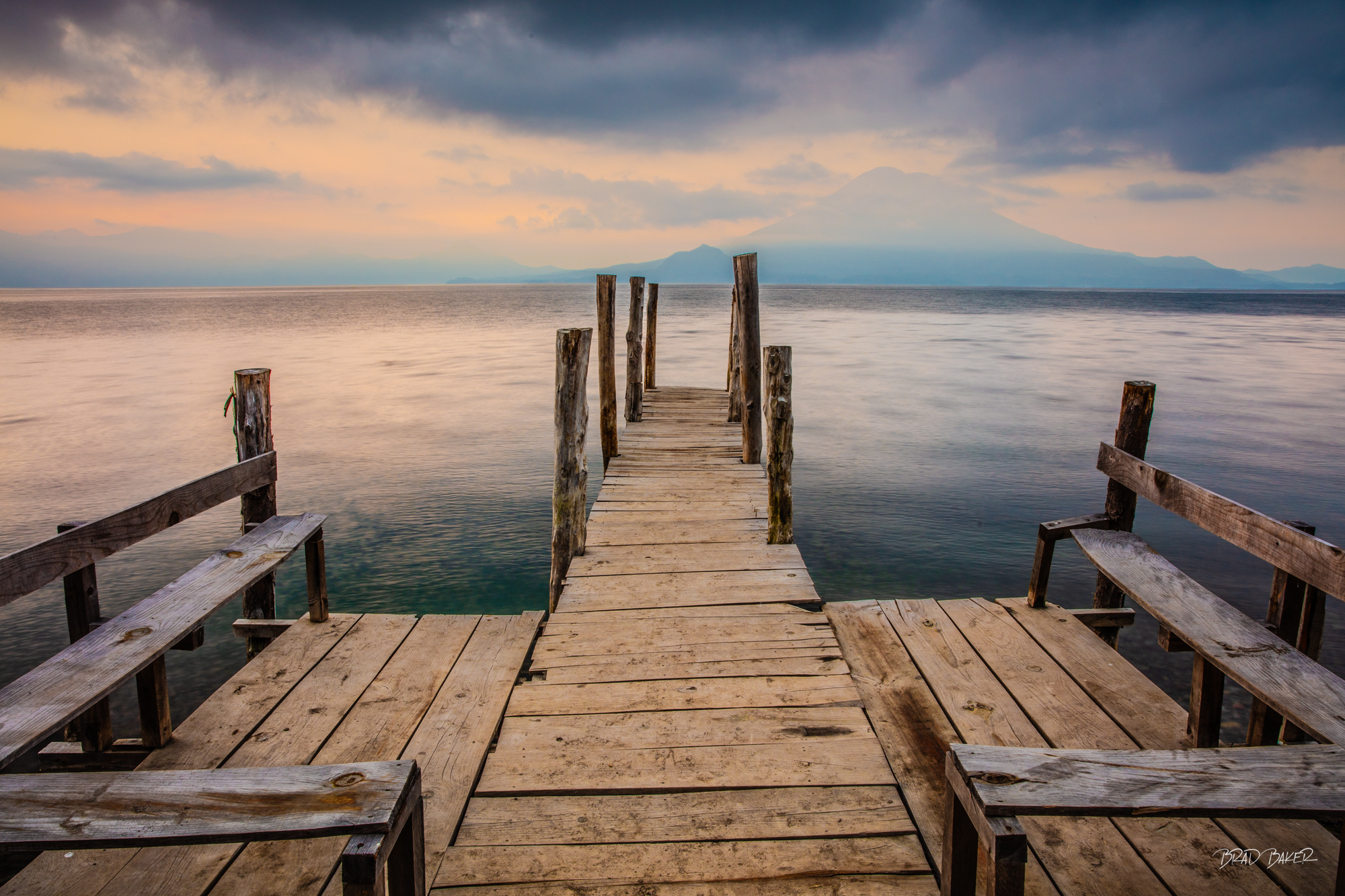 A place to relax Weathered wooden dock with rustic poles extends over calm lake, volcanic peak rising in background, Lake Atitlan Guatemala